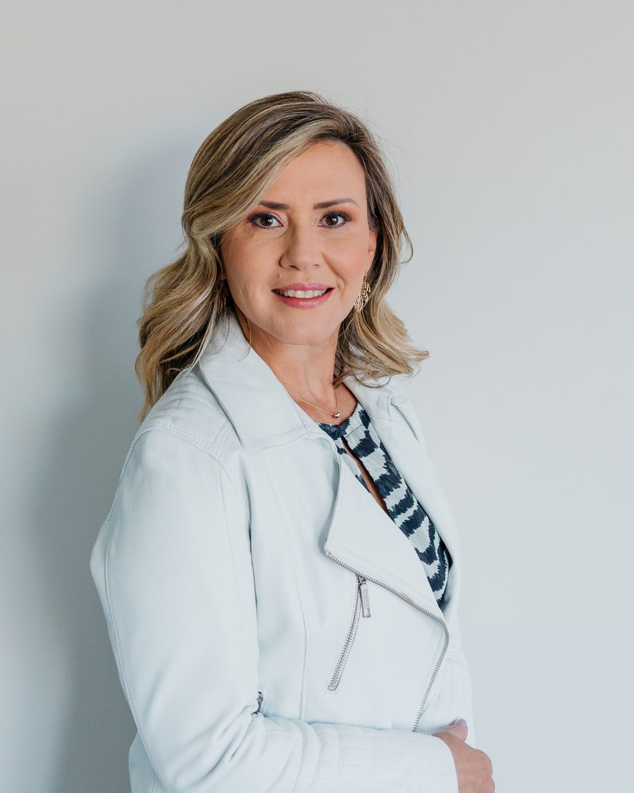 Gabi Caporali, consultora de atendimento, de jaqueta branca, diante de uma parede clara.